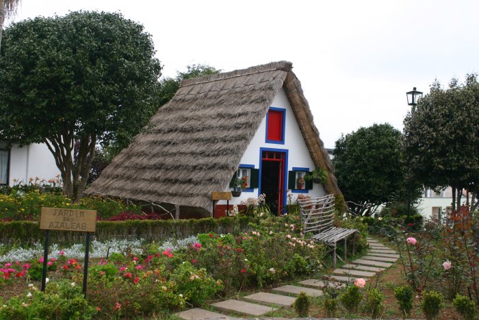 Traditionellt hus med halmtak, vit fasad, blå fönsterluckor och dörr, omgivet av en trädgård med blommande azaleor.