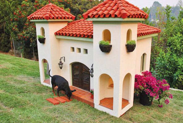 En stor hund utforskar entrén till ett lyxigt hundhus som ser ut som ett litet slott med rött tegeltak.