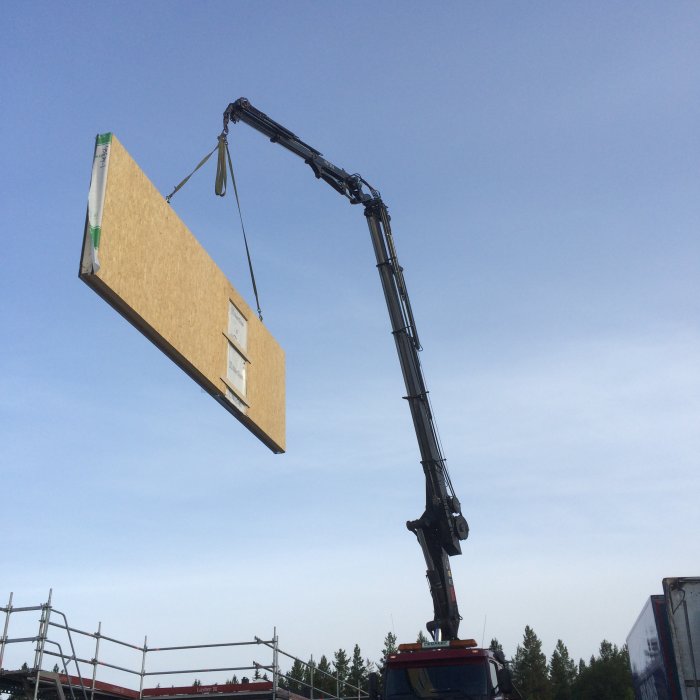 Väggsektion med fönster lyfts av kran vid en husstomresning mot en blå himmel.