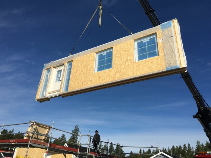 Väggsektion med fönster hissas av kran vid husstomresning mot blå himmel.