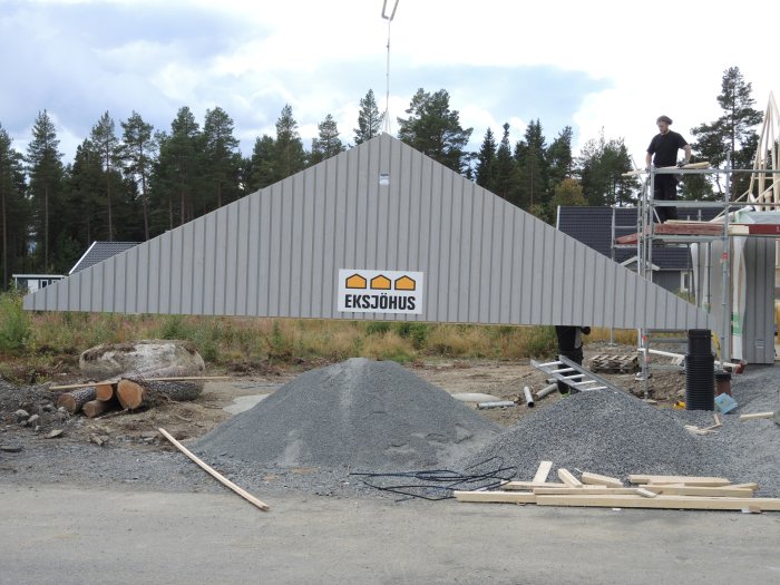 Byggarbetsplats med husvägg som hissas och byggarbetare, högar av grus och skog i bakgrunden.