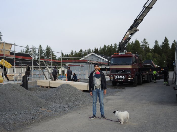 Person med hund framför byggarbetsplats där en husstomme reses med hjälp av en kranbil.