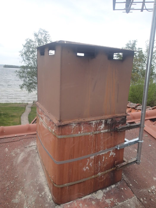Skorsten med avtagbar topp-huv och vittrad murning på ett tak, med utsikt över sjö.