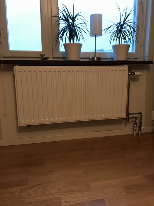 Nyinstallerad radiator under fönster med synliga rör och äldre termostat, inte centrerad och nära fönsterbräda.