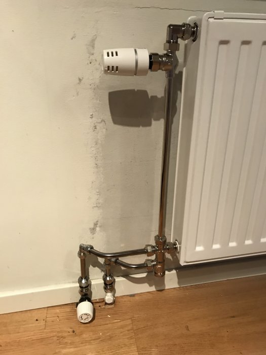 Nyinstallerad radiator där rör leder upp till termostat, gamla termostatventilen kvar vid golv, slarvig väggfinish.