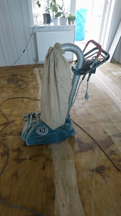 Golvslipmaskin på partiellt slipat trägolv med slipdammsäck och elektrisk kabel.