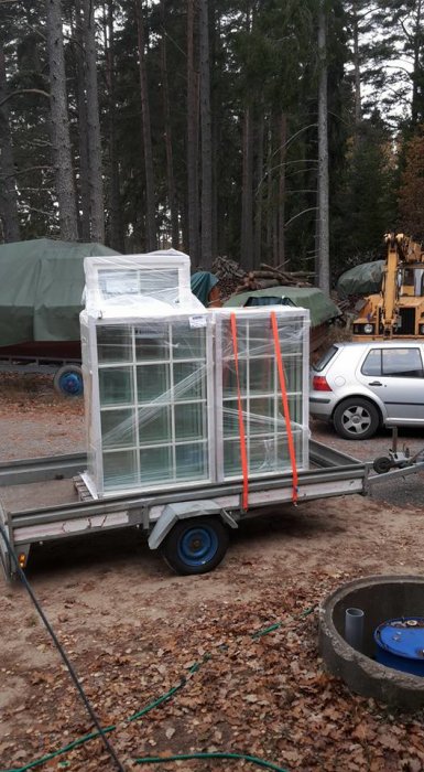 Nya fönster från Elit fönster inslagna i plast och säkrade med orange band på en släpvagn i skogsmiljö.