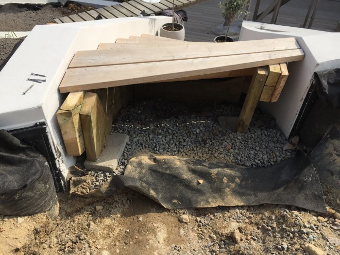 En träbänk under konstruktion med stöd av klumpiga träklossar och skruvar på en grusig mark.