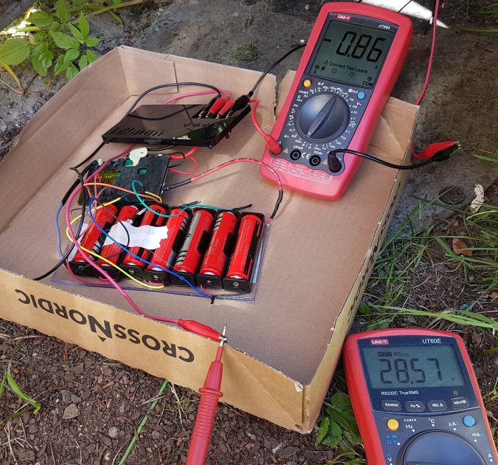 En hemmagjord batteripack med röda och svarta 18650-batterier, anslutna med multicolorkablar, testas med två multimetrar.