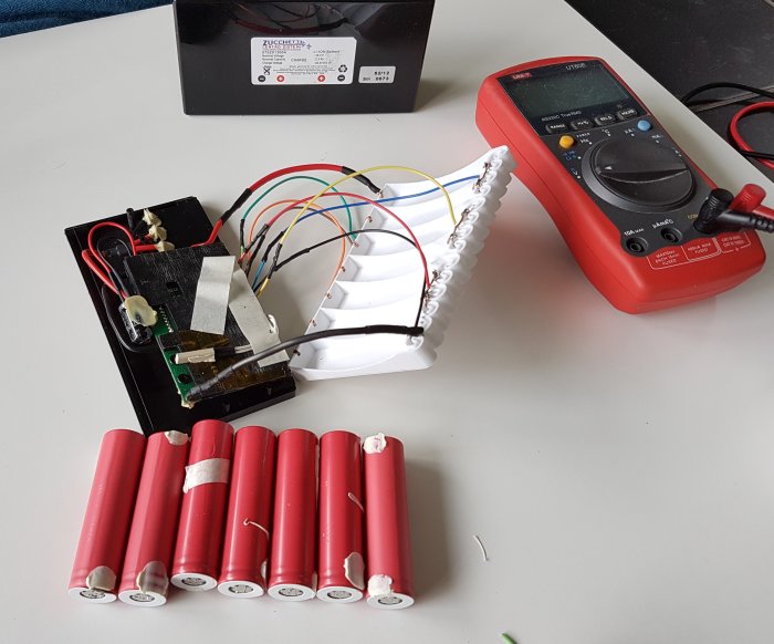 Elektronikkomponenter och röda 18650-battericeller på bord, bredvid multimeter och batteriladdare.