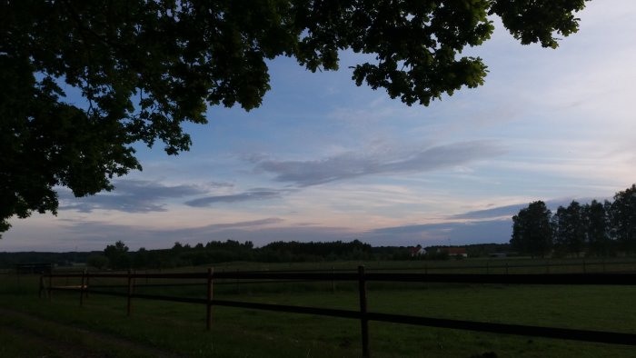 Kvällshimmel med träd och ett staket i förgrunden på en lantlig tomt.