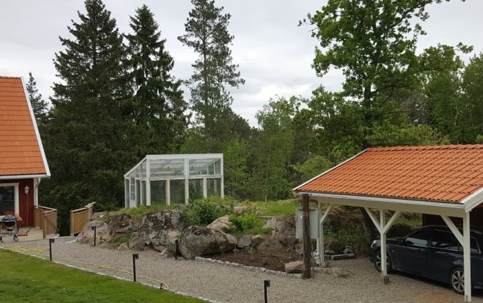 Trädgård med växthus, omgärdad av stenar och berg, intill en carport med insynsskydd.