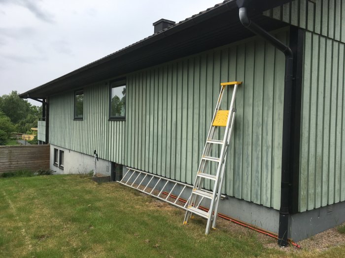 Sliten fasad på husets östsida med en stege lutad mot väggen och avkopplade spottar vid takfoten.