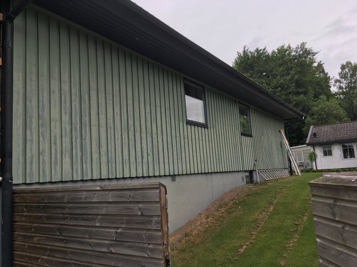 Sliten grön fasad på östsida av hus med början av renoveringsarbete, stegar lutade mot väggen.