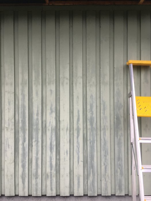 Sliten fasadpanel på öst-sidan av ett hus med en stege lutad mot väggen.