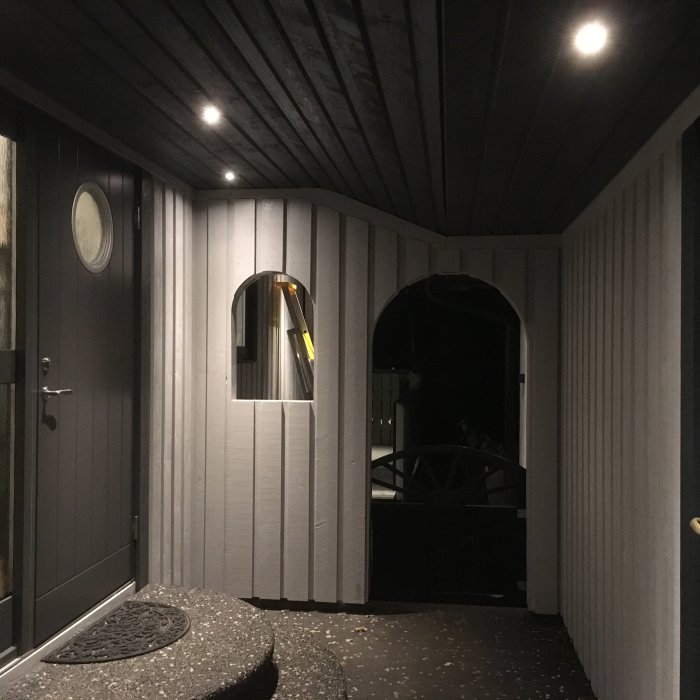 Upplyst veranda med mörkgrå panel och vit struktur, öppen svart dörr och en vägglykta.