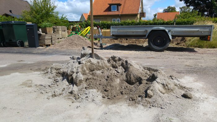 Hög med stenmjöl mitt i en gata med spade, släpvagn och omgivande byggmaterial i bakgrunden.