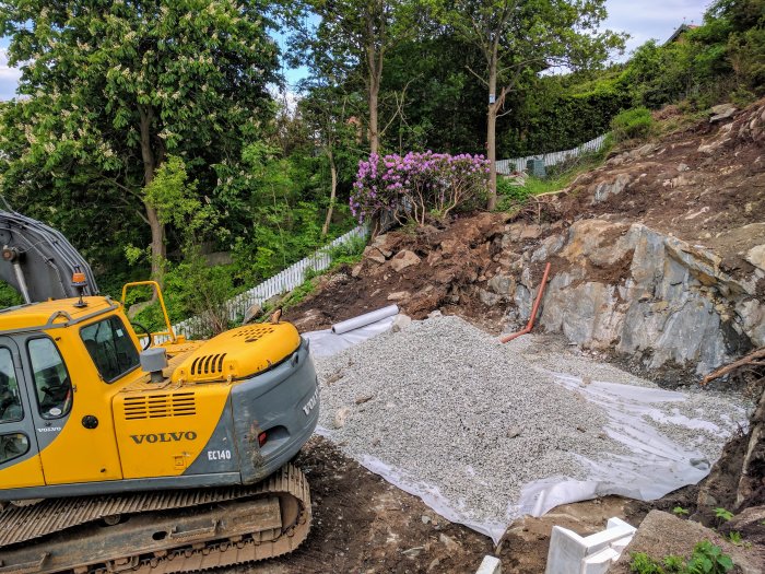 En grävmaskin från Volvo vid en byggarbetsplats med grushög och utsprängd klippa i en trädgård.