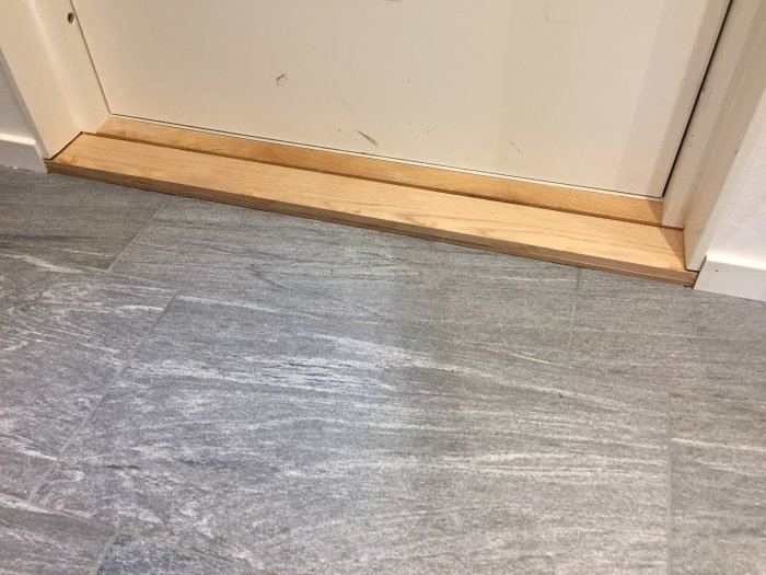 Nyinstallerad trätröskel i ek vid dörren i en hall med grått klinkergolv, arbetet ej helt slutfört.