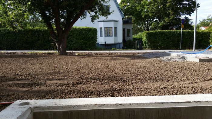 Nyfräst jord i förgrunden med betongfundament, hus och häck i bakgrunden samt en blå slang.