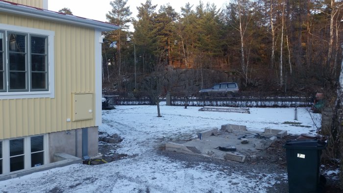 Husets hörn med utbyggd del, källartrappa, snötäckt gräsmatta och nedmonterat förråd.