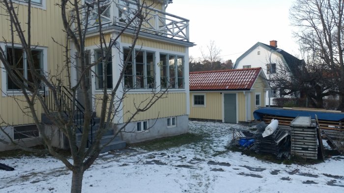 Utbyggnad av hus med ny källartrappa och förråd, lätt snötäckt gräsmatta framför och materialhögar till höger.