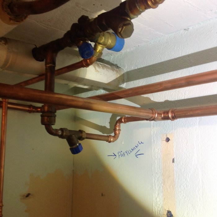 Rörinstallation med kopparledningar och en blå balkongventil, markerad med en handskriven grön pil på väggen.