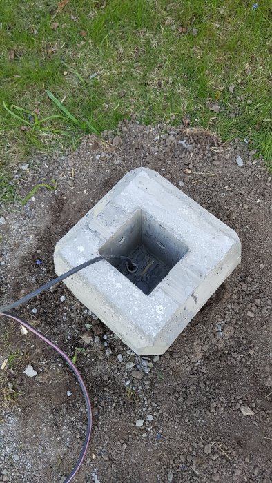 Nyfärdigt, kvadratiskt betongfundament i marken med öppning i mitten och en kabel genomdragen.