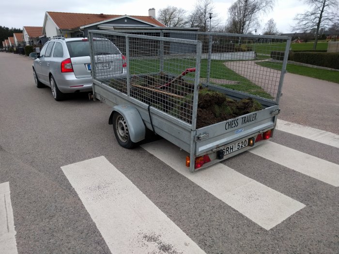 Släpvagn full med grävd lera parkerad på en gata bakom en silverfärgad bil.