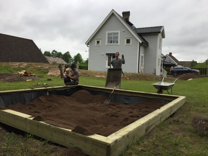 Nybyggd lekplats med sandlåda, personer arbetar med skottkärror, hus i bakgrunden.