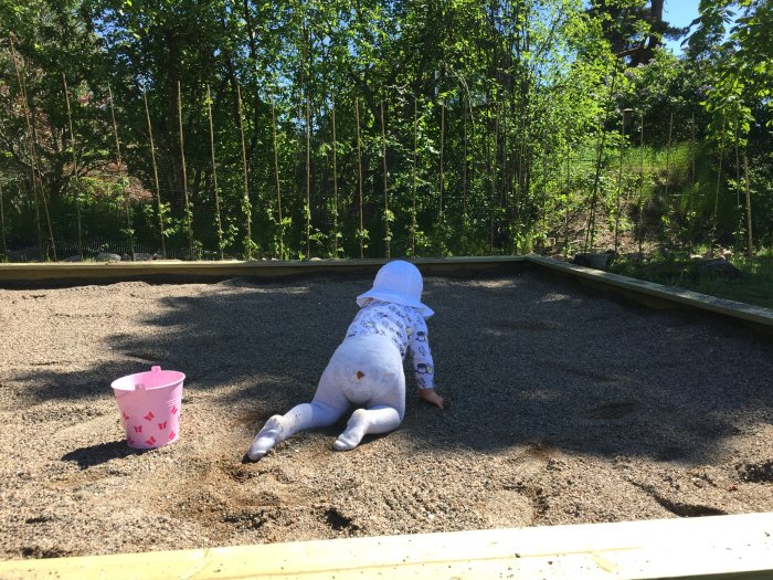 Barn leker i en nybyggd sandlåda på en solig tomt med grönska i bakgrunden.