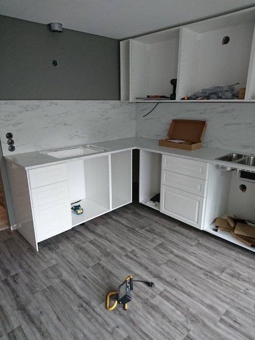 Kök under renovering med vit marmormönstrad bänkskiva, vita skåpstommar och verktyg på golvet.