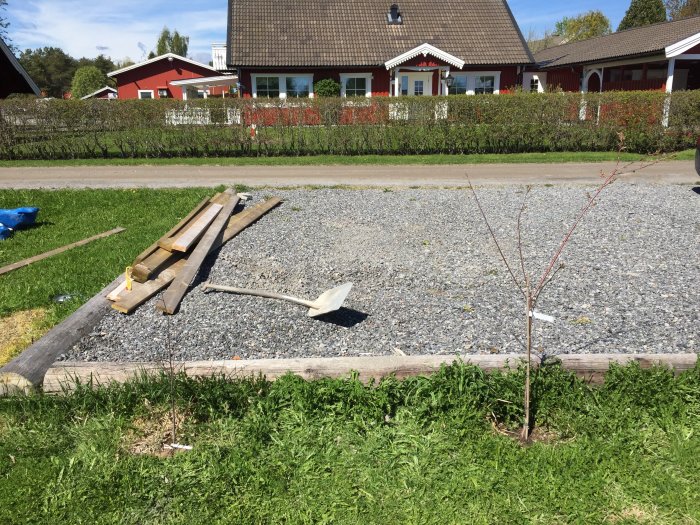 Nyplanterat träd och spade vid nygrusad yta med timmerkant framför rött hus.