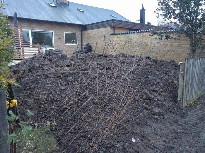 Stor jordhög och grovavjämnade massor på en bostadstomt med bakgrund av hus och häck.