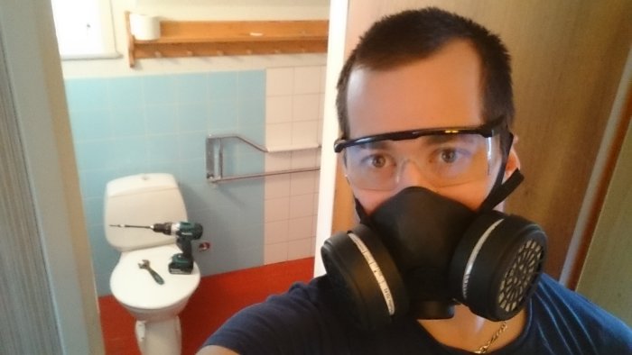 Person med skyddsmask tar selfie i badrum med ljusblå kakel innan renovering.