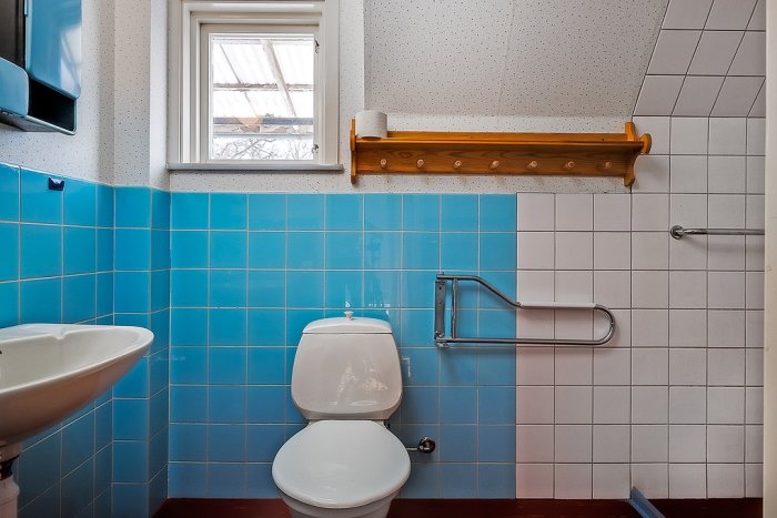 Ett gammalt badrum med blå kakel halvvägs upp på väggarna, vit toalett och handfat, samt stycke av ett badkars kant.