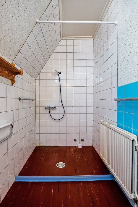 Före renovering, ett badrum från -64 med vita kakelväggar, mörkt trägolv och dusch hörna, speglande en tidstypisk stil.