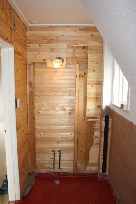 Rivningsfas i badrum med träväggar, synliga rörinstallationer och röd golvmatta.