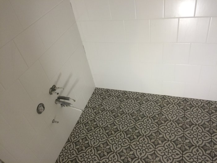 Renoverat badrum med vita väggkakel och mönstrade matta klinkers, installationer för vatten ej färdigställda.