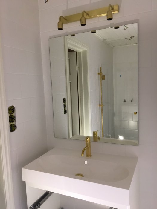 Renoverat badrum med vit handfat, guld kran och armaturer, spegel, och synlig dusch bakom.