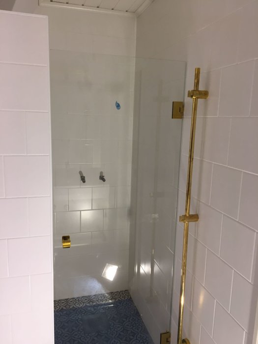 Nyrenoverad duschhörna med vita kakelväggar, guldiga duschdetaljer och blått mönstrat golv.