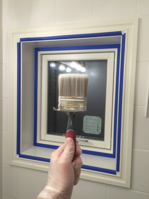 Person målar karmen runt ett nyligen installerat fönster som är maskerat med blå tejp.