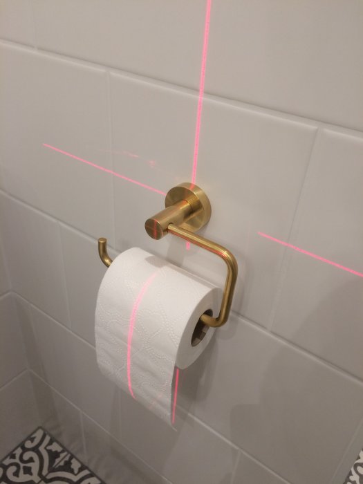 Gyllene toalettpappershållare med en rulle papper, monterad på en vit kakelvägg, med en rosa laserlinje för noggrann positionering.