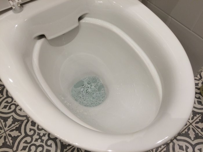 Ifö Spira toalett utan spolkant med kristallblått vatten och mönstrade golvplattor.