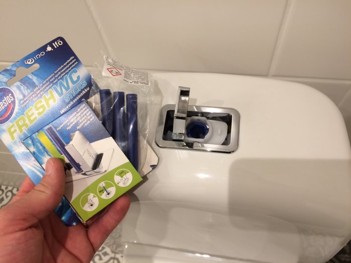 Hand som håller en förpackning med Fresh WC-sticks framför en Ifö Spira-toalett med öppen spolknapp och blått vatten.