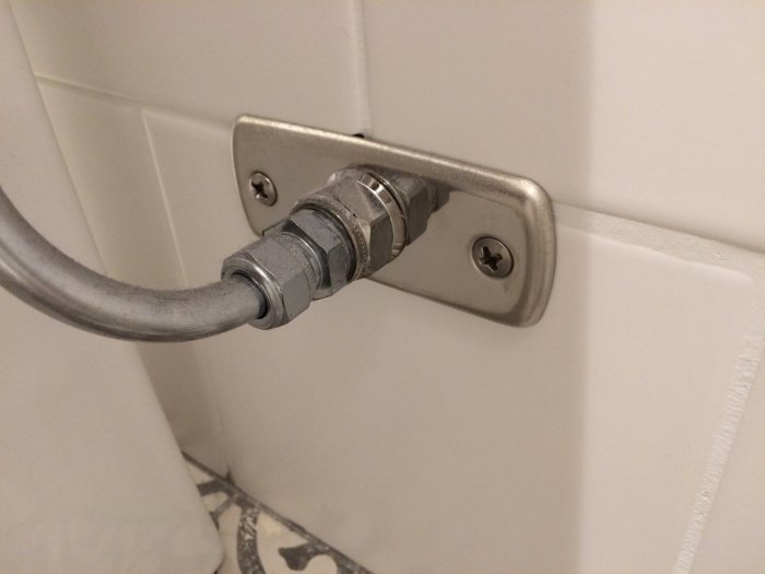 Mässingsdetalj på handdukstork i badrum med vita kakelväggar och gråa rör.