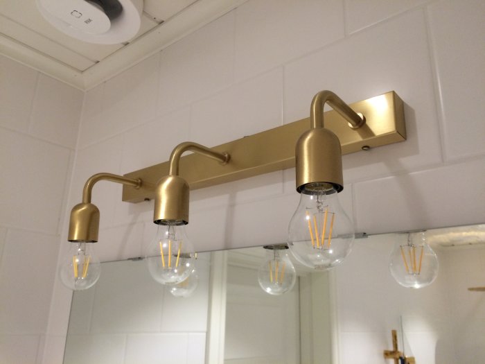 Mässingslampa med tre glödlampor på vit kakelvägg i badrum, avspeglar ägarens nöjda val av material.