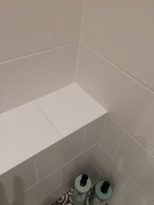 Välvinklade vita kakelväggar i ett badrumshörn med en hylla och två flaskor på golvet.