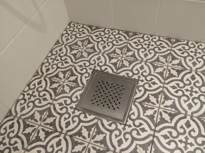 Mönstrade klinkergolv i badrum med en elegant golvbrunn och vita kakelväggar i bakgrunden.