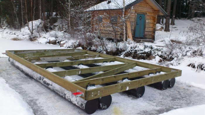 En flotte under konstruktion med träreglar och svarta cylindriska pontoner ligger på en isbelagd yta, omgiven av snö och en liten stuga i bakgrunden.